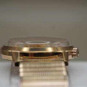 Op de afbeelding: Een goudkleurig polshorloge met een gepolijste metalen kast en een bijpassende fijnmazige metalen band. Het horloge heeft een opvallende, licht gewelfde kristallen wijzerplaat en een kleine, geribbelde opwindkroon aan de zijkant van de ronde kast.