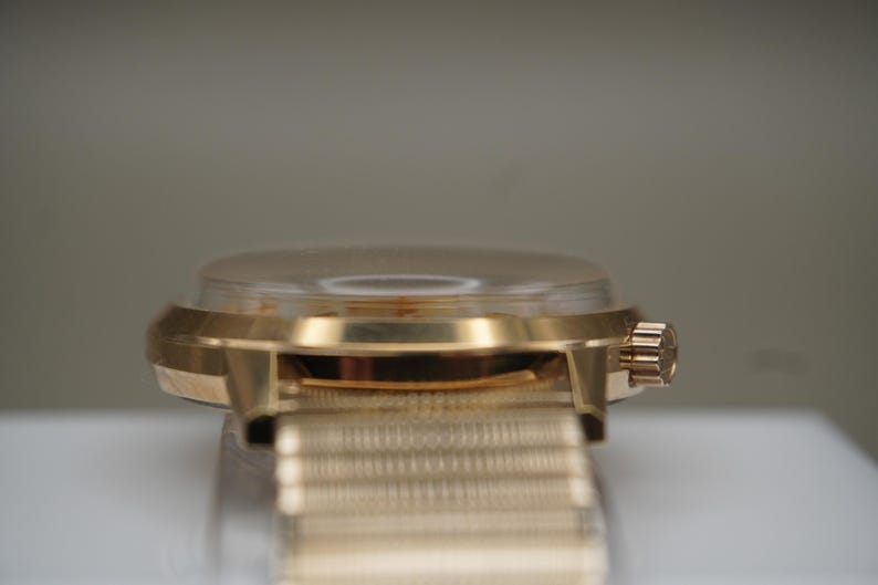 Op de afbeelding: Een goudkleurig polshorloge met een gepolijste metalen kast en een bijpassende fijnmazige metalen band. Het horloge heeft een opvallende, licht gewelfde kristallen wijzerplaat en een kleine, geribbelde opwindkroon aan de zijkant van de ronde kast.