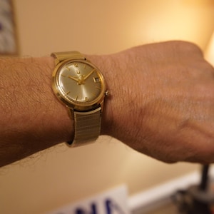 Op de afbeelding: Een goudkleurig analoog polshorloge met een ronde champagnekleurige wijzerplaat met goudkleurige uurmarkeringen, slanke wijzers en een datumvenster op de drie-uurpositie. Het horloge is voorzien van een flexibele, gestructureerde metalen rekband.
