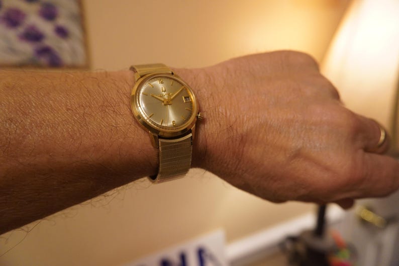 Op de afbeelding: Een goudkleurig analoog polshorloge met een ronde champagnekleurige wijzerplaat met goudkleurige uurmarkeringen, slanke wijzers en een datumvenster op de drie-uurpositie. Het horloge is voorzien van een flexibele, gestructureerde metalen rekband.