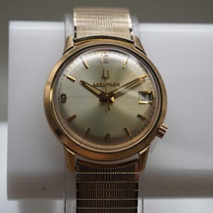 Op de afbeelding: Een goudkleurig analoog polshorloge met een ronde champagnekleurige wijzerplaat met de tekst Accutron, een datumvenster op de drie-uurpositie, goudkleurige uurmarkeringen en een bijpassende goudkleurige metalen rekband met textuur.