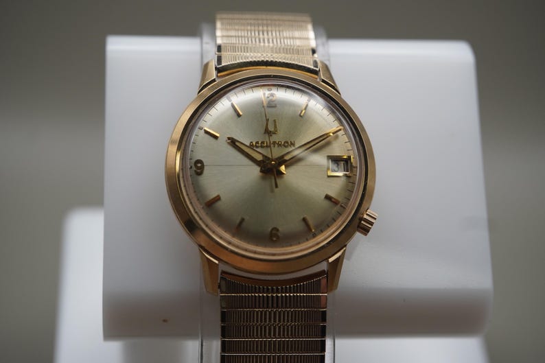 Op de afbeelding: Een goudkleurig analoog polshorloge met een ronde champagnekleurige wijzerplaat met de tekst Accutron, een datumvenster op de drie-uurpositie, goudkleurige uurmarkeringen en een bijpassende goudkleurige metalen rekband met textuur.