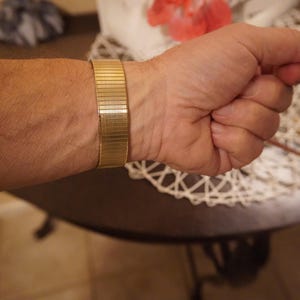 Op de afbeelding: Een goudkleurige metalen rekbare horlogeband met een fijne horizontale geribbelde textuur. De band is opgebouwd uit meerdere in elkaar grijpende rechthoekige schakels die een flexibel, doorlopend lusontwerp vormen met een gepolijste metalen afwerking.
