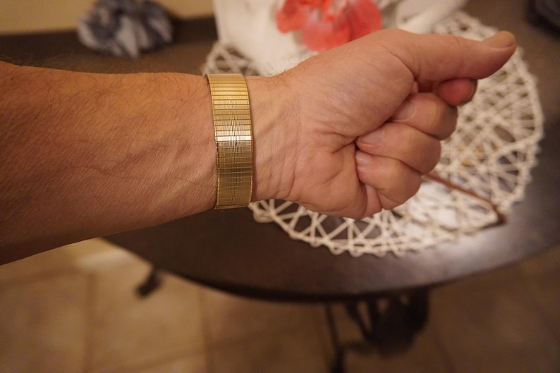 Op de afbeelding: Een goudkleurige metalen rekbare horlogeband met een fijne horizontale geribbelde textuur. De band is opgebouwd uit meerdere in elkaar grijpende rechthoekige schakels die een flexibel, doorlopend lusontwerp vormen met een gepolijste metalen afwerking.