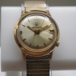 Op de afbeelding: Een goudkleurig analoog polshorloge met een ronde champagnekleurige wijzerplaat, voorzien van het Accutron-logo, goudkleurige wijzers en uurmarkeringen, een datumvenster op de drie-uurpositie en een flexibele goudkleurige metalen horlogeband.