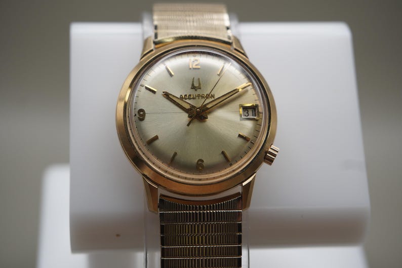 Op de afbeelding: Een goudkleurig analoog polshorloge met een ronde champagnekleurige wijzerplaat, voorzien van het Accutron-logo, goudkleurige wijzers en uurmarkeringen, een datumvenster op de drie-uurpositie en een flexibele goudkleurige metalen horlogeband.