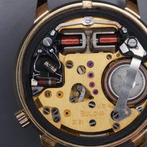Op de afbeelding: Het interne mechanisme van een goudkleurig Bulova-horloge met het modelnummer 2181 zichtbaar op de plaat. Het uurwerk bevat een rond batterijcompartiment, koperdraadspoelen en diverse metalen tandwielen en elektronische componenten.