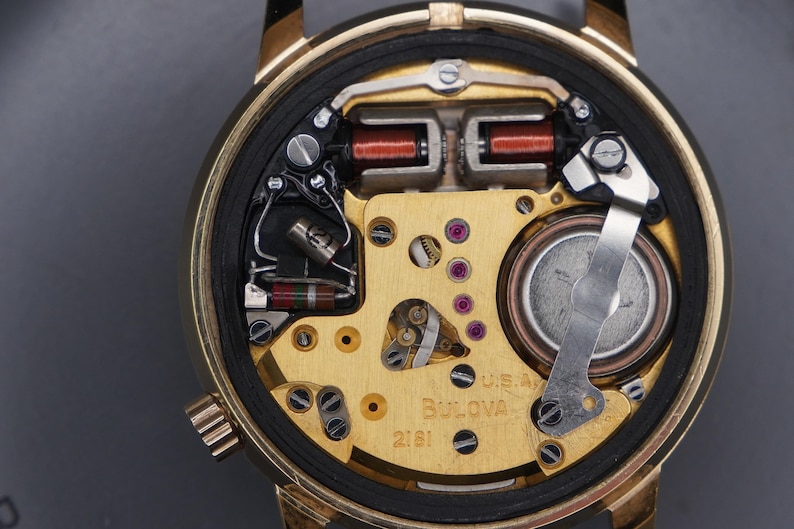 Op de afbeelding: Het interne mechanisme van een goudkleurig Bulova-horloge met het modelnummer 2181 zichtbaar op de plaat. Het uurwerk bevat een rond batterijcompartiment, koperdraadspoelen en diverse metalen tandwielen en elektronische componenten.