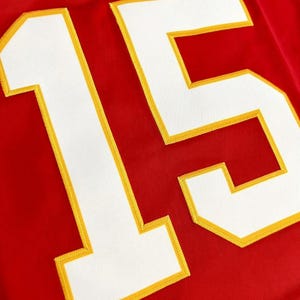 Peut inclure: Un maillot rouge vif arborant le num&eacute;ro 15 en grands chiffres blancs. Les chiffres sont soulign&eacute;s par une &eacute;paisse bordure jaune et sont cousus sur le tissu avec une texture visible, cr&eacute;ant un contraste marqu&eacute; sur le fond rouge uni.
