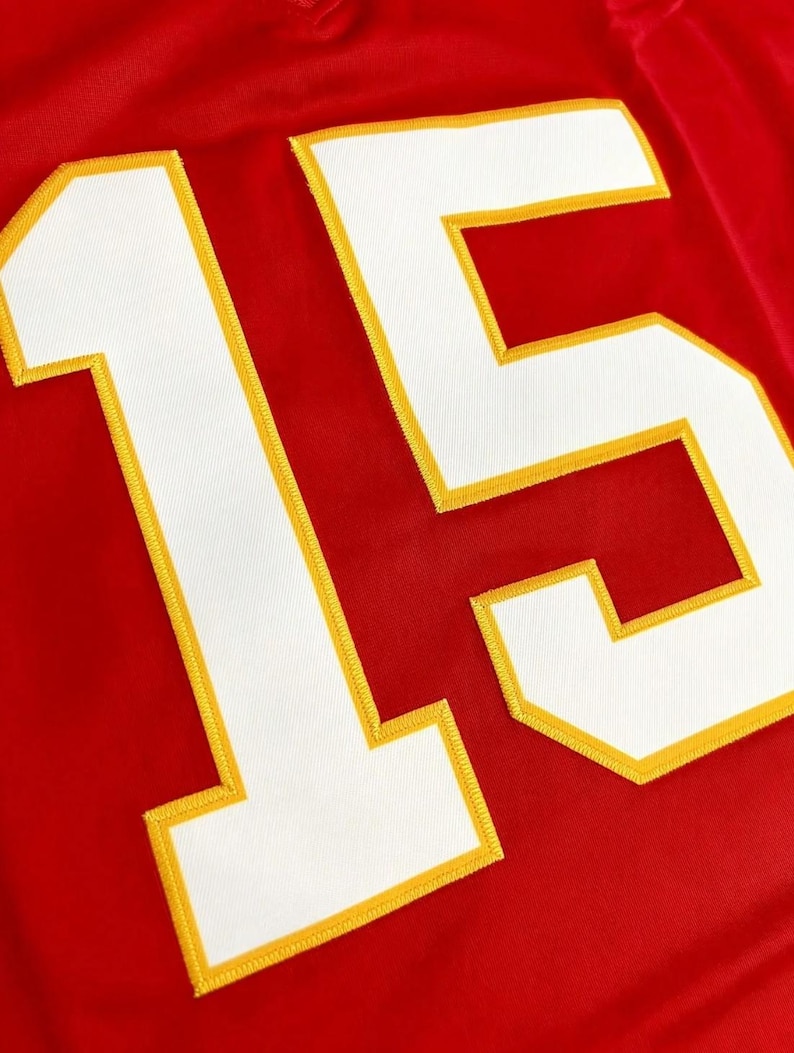 Peut inclure: Un maillot rouge vif arborant le num&eacute;ro 15 en grands chiffres blancs. Les chiffres sont soulign&eacute;s par une &eacute;paisse bordure jaune et sont cousus sur le tissu avec une texture visible, cr&eacute;ant un contraste marqu&eacute; sur le fond rouge uni.