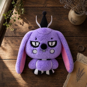 Puede incluir: Un peluche de una criatura morada y peluda con orejas largas, un sombrero negro puntiagudo y detalles en blanco. El personaje tiene ojos grandes con pupilas negras, una nariz pequeña negra, dos dientes blancos y mejillas redondas blancas.