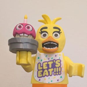 May include: A large yellow plastic figure resembling a chicken with a white bib that says LET'S EAT!!! and a pink cupcake accessory, standing behind a small silver and black armored warrior figure holding a gray spear.