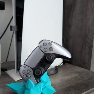 Könnte beinhalten: Ein grauer und schwarzer Videospiel-Controller ruht auf einem türkisfarbenen geometrischen Ständer. Im Hintergrund befindet sich eine weiße und schwarze Spielkonsole. Der Controller hat silberne Akzente und schwarze Tasten.