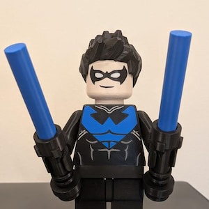 May include: A large, 3D-printed Nightwing action figure with black hair, a black domino mask, and a black suit featuring a blue chest emblem, holding two blue light-up sticks. A small, standard-sized blonde minifigure stands at its feet.