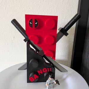 Puede incluir: Un soporte de exhibición estilo bloque rojo y negro con un emblema de máscara de Deadpool, dos espadas grises cruzadas y el texto BANG! en letras rojas. Una pequeña figura de plástico gris y negra se encuentra frente a la estructura sobre una superficie blanca.
