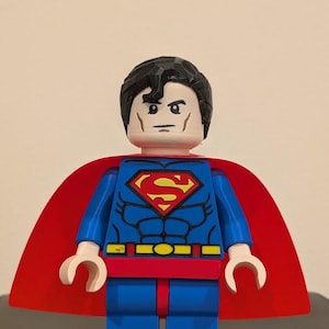 May include: A Superman minifigure featuring a blue torso with a red and yellow chest emblem, black hair, and a red cape. The figure has white skin, blue legs, and white hands. The chest design includes black line details representing muscle definition.