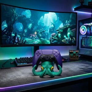 Könnte beinhalten: Gaming-Setup mit einem gebogenen Monitor, der eine Unterwasserszene zeigt. Ein lila Controller liegt auf einem Oktopus-Ständer. Ein Gaming-PC mit bunten Lichtern und ein Headset vervollständigen das Set. Eine Tastatur und ein Mauspad mit leuchtendem Rand sind ebenfalls vorhanden.