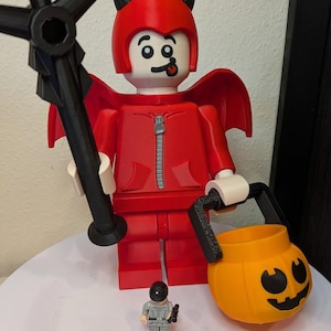 May include: A large red devil figure with black horns, a zip detail on the chest, and red wings, holding a black trident and an orange jack-o'-lantern bucket. A small grey minifigure stands in front of the large figure on a white surface.