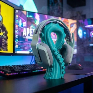 Puede incluir: Un soporte para auriculares con forma de pulpo turquesa sostiene unos auriculares para juegos blancos y grises. El soporte está sobre un escritorio negro frente a dos monitores de ordenador que muestran gráficos de videojuegos. También se ven un teclado y una torre de ordenador.