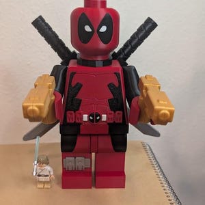 May include: A large red and black plastic building block figure resembling Deadpool, featuring two gold-coloured toy pistols in its hands and two black swords strapped to its back. Beside it stands a small, standard-sized building block figure.