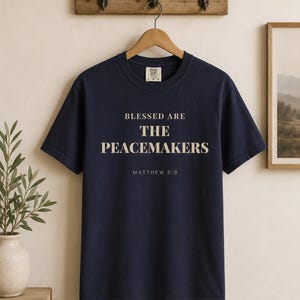 Op de afbeelding: Marineblauwe t-shirt met de tekst "BLESSED ARE THE PEACEMAKERS" en "MATTHEW 5:9" in lichtbeige. Het shirt hangt aan een houten hanger tegen een neutrale muur.