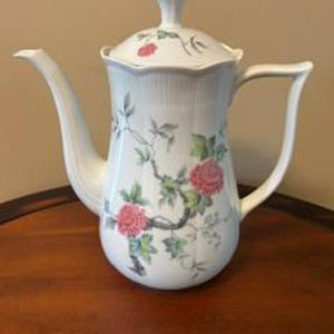 May include: A white porcelain teapot featuring a scalloped rim and a lid topped with a gold-colored finial. The body is decorated with a delicate botanical pattern of pink flowers, green leaves, and dark brown branches, complemented by a curved handle.