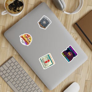 Może przedstawiać: Kolekcja czterech naklejek z motywami inżynierii oprogramowania. Naklejki zawierają teksty takie jak "Software Engineer" i "Bug Life is Eternal". Naklejki znajdują się na szarym laptopie.