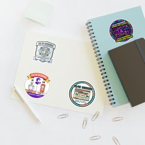 May include: A collection of stickers and a notebook. The stickers feature designs with the text "UX/UI DESIGNER" and related graphics. One sticker has the text "USERS ARE CHAOS". The notebook is light blue with a dark brown cover. Paper clips and a pen are also present.