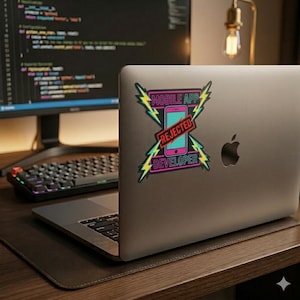May include: A laptop with a sticker that reads "MOBILE APP REJECTED DEVELOPER." The sticker features a pink smartphone with a teal screen, crossed by a red banner. Yellow lightning bolts frame the text, with a black keyboard and monitor in the background.