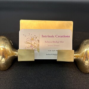 May include: A brass business card holder with a unique design. The holder features two rounded end pieces connected by a rectangular base, with a card displaying the text "Intrinsic Creations". The card has a floral design and contact information.