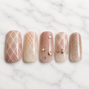 May include: Five artificial nails with a marble background. The nails feature various designs, including a pink and white lattice pattern, glitter, and heart-shaped and gem embellishments. The nails are in shades of pink, peach, and gold.