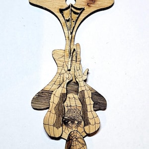 May include: A laser-cut wooden ornament featuring a detailed engraving of a superhero character hanging upside down by a web. The design includes a web pattern on the character's suit and a circular hole at the top for hanging purposes.