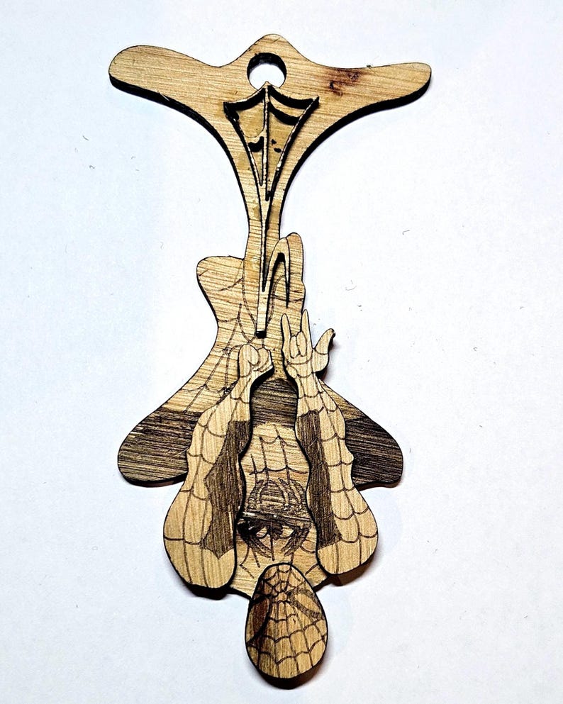 May include: A laser-cut wooden ornament featuring a detailed engraving of a superhero character hanging upside down by a web. The design includes a web pattern on the character's suit and a circular hole at the top for hanging purposes.