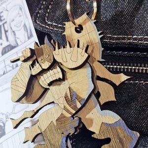 May include: A laser-cut wooden keychain featuring a stylized figure of a caped character with spiked shoulder armor and a mask. The light-colored wood has dark engraved lines and subtle blue-tinted accents, attached to a gold-toned metal ring.