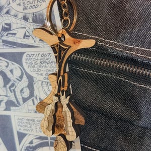 May include: A laser-cut wooden keychain featuring a stylized figure hanging upside down with a web pattern, attached to a gold-toned metal split ring and lobster clasp. The wood has natural grain variations and dark etched details throughout the design.