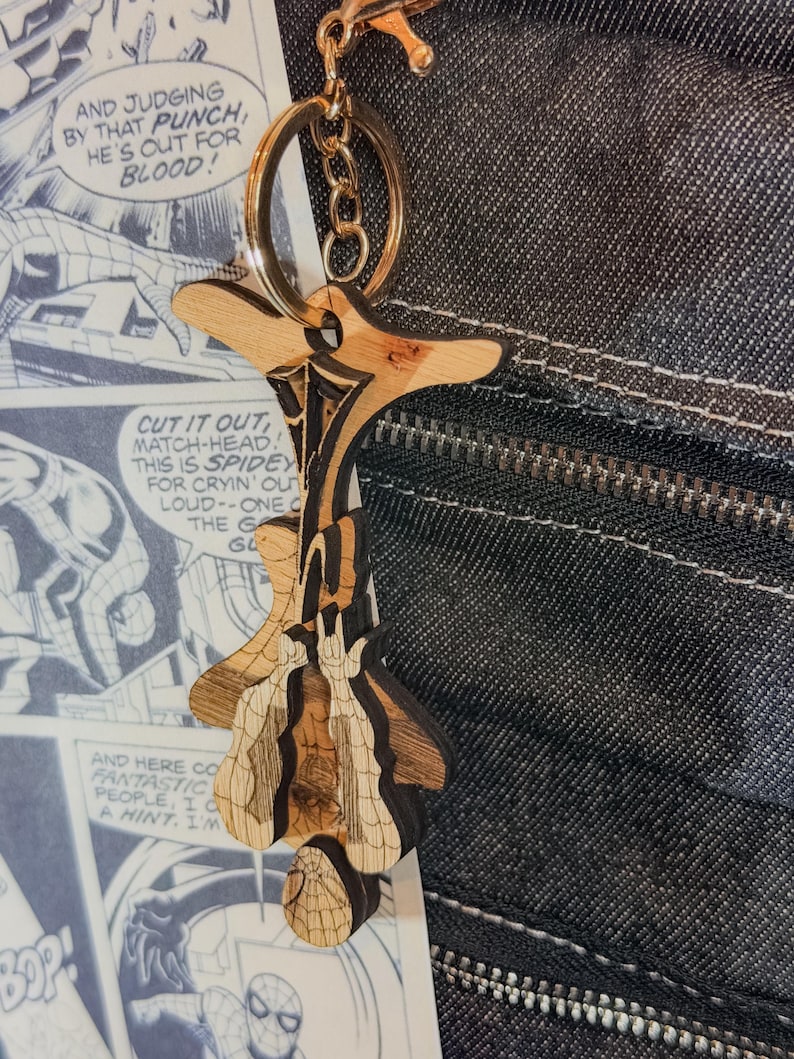 May include: A laser-cut wooden keychain featuring a stylized figure hanging upside down with a web pattern, attached to a gold-toned metal split ring and lobster clasp. The wood has natural grain variations and dark etched details throughout the design.