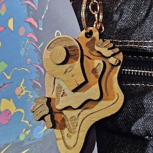 May include: A layered wooden keychain featuring an abstract, organic shape with a circular top element and a gold-toned metal ring attachment. The wood displays a natural grain pattern with darker, etched lines defining the contours of the design.