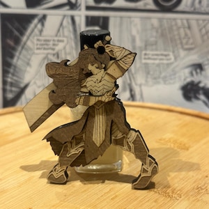 May include: A laser-cut wooden ornament featuring a stylized warrior figure with long flowing hair, wearing armor and holding a large rectangular sword. The figure is depicted in a dynamic combat stance with intricate engraved details on the wood surface.