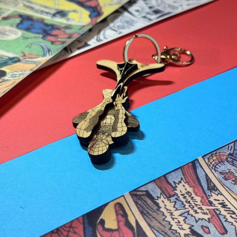 May include: A layered wooden keychain featuring a laser-cut design of a superhero character in a classic web-slinging pose. The keychain includes a gold-toned metal split ring and a lobster clasp, with intricate black etched details on the light wood.