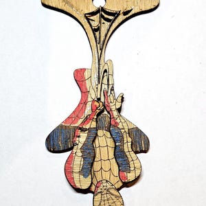 May include: A laser-cut wooden ornament featuring a hanging superhero character in a red and blue suit with a web pattern. The figure is suspended from a top section with a circular hole for hanging, showcasing natural wood grain and etched details.