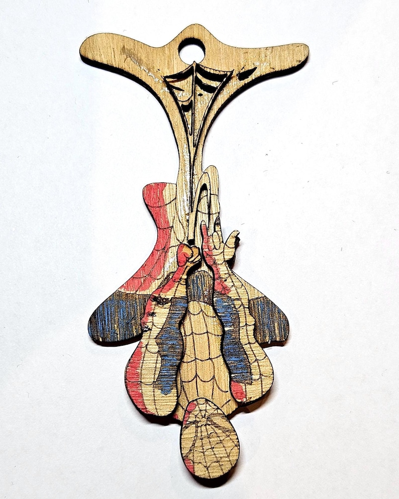 May include: A laser-cut wooden ornament featuring a hanging superhero character in a red and blue suit with a web pattern. The figure is suspended from a top section with a circular hole for hanging, showcasing natural wood grain and etched details.