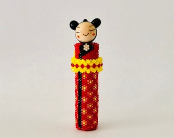 May include: A small, beaded doll wearing a red and yellow patterned dress. The doll has a black and white face with a smiling expression.