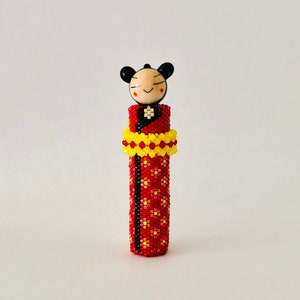 May include: A small, beaded doll wearing a red and yellow patterned dress. The doll has a black and white face with a smiling expression.