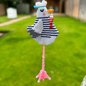 May include: A handmade seagull ornament with a sailor hat and striped shirt. The seagull is holding a red and white fish in its beak. The ornament has a pink leg and foot, and is hanging from a natural twine.