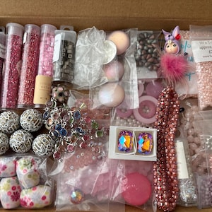 May include: A box overflowing with craft supplies, featuring beads, rhinestones, and decorative elements in pink, purple, and clear hues. The selection includes glass beads, round beads, and various embellishments.