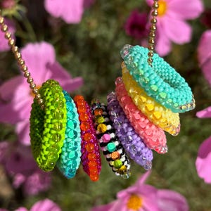 Puede incluir: Un collar de cuentas de colores con una pila de anillos redondos de cuentas en tonos de verde, turquesa, rosa, morado, amarillo y naranja. El collar está colgado de una cadena dorada.