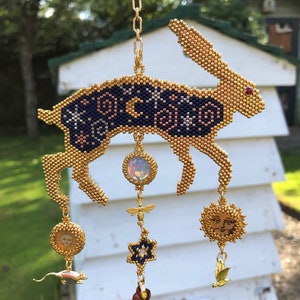 May include: A gold beaded rabbit with a blue and gold patterned body hangs from a gold chain. The rabbit has a red gemstone eye and is adorned with dangling charms, including a sun, a star, a dragonfly, a mouse, and a heart.
