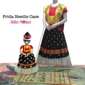 Puede incluir: Un estuche para agujas de cuentas con forma de mu&ntilde;eca Frida Kahlo. La mu&ntilde;eca lleva un vestido rojo, amarillo y negro con un borde de encaje blanco. La cara de la mu&ntilde;eca est&aacute; pintada con un dise&ntilde;o de calavera de az&uacute;car. El estuche para agujas est&aacute; sobre un libro titulado "Frida Kahlo: Un retrato &iacute;ntimo".