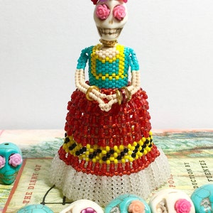 Puede incluir: Una mu&ntilde;eca de cuentas con cara de calavera, que lleva un top de cuentas turquesa y amarillo, una falda de cuentas roja y un sombrero de cuentas negro con flores rosas y rojas. La mu&ntilde;eca est&aacute; de pie sobre un libro con un collar de calavera turquesa, blanco y rosa.