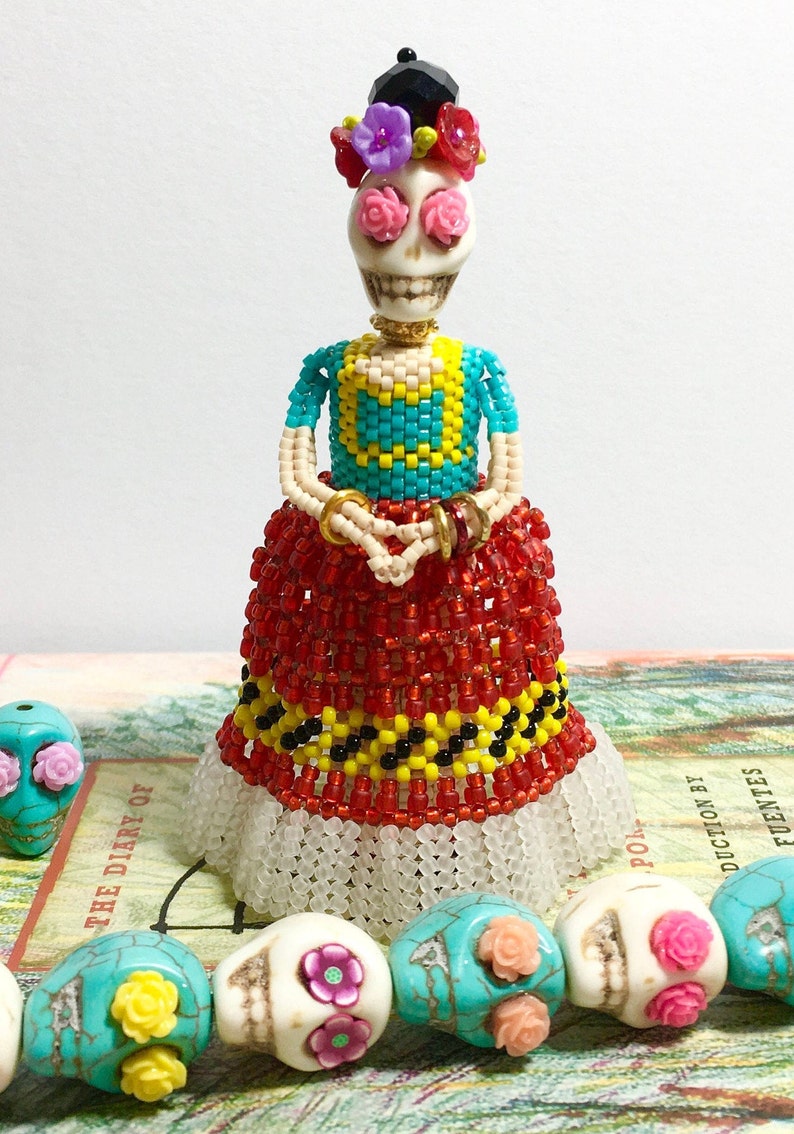 Puede incluir: Una mu&ntilde;eca de cuentas con cara de calavera, que lleva un top de cuentas turquesa y amarillo, una falda de cuentas roja y un sombrero de cuentas negro con flores rosas y rojas. La mu&ntilde;eca est&aacute; de pie sobre un libro con un collar de calavera turquesa, blanco y rosa.
